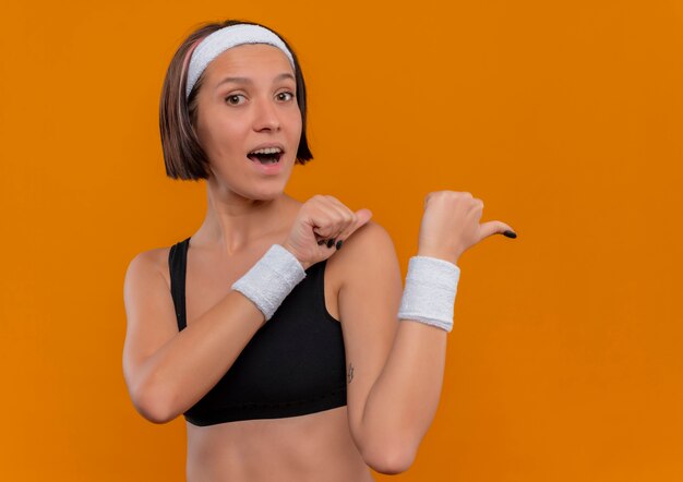 Young fitness woman in sportswear with headband positive and happy pointing back with thumbs standing over orange wall