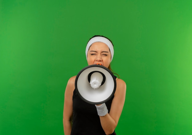 Young fitness woman in sportswear with headband shouting to megaphone with aggressive expression standing over green wall