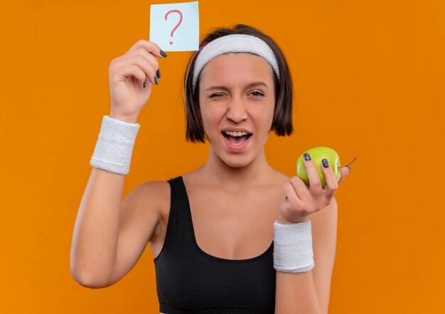 Young fitness woman in sportswear with headband showing reminder paper with question mark holding green apple winking and smiling with happy face standing over orange wall