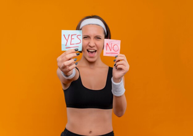 Молодая фитнес-женщина в спортивной одежде с повязкой на голову показывает два напоминания со словом да и нет, весело улыбаясь, стоя над оранжевой стеной