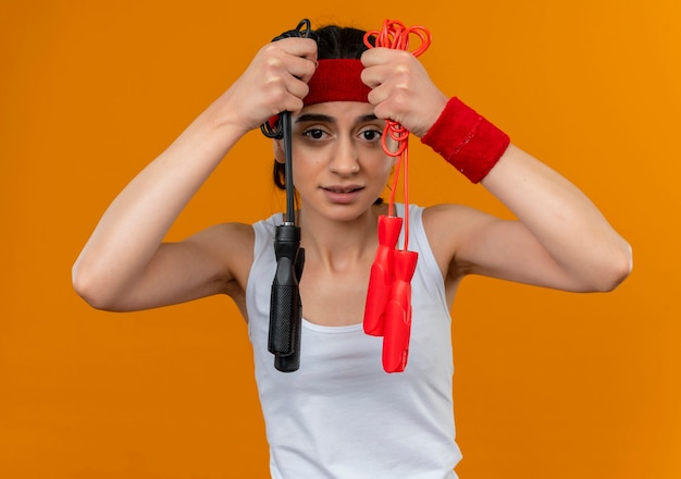 Foto gratuita giovane donna fitness in abbigliamento sportivo con fascia che mostra due corde per saltare cercando confuso avendo dubbi in piedi sopra la parete arancione