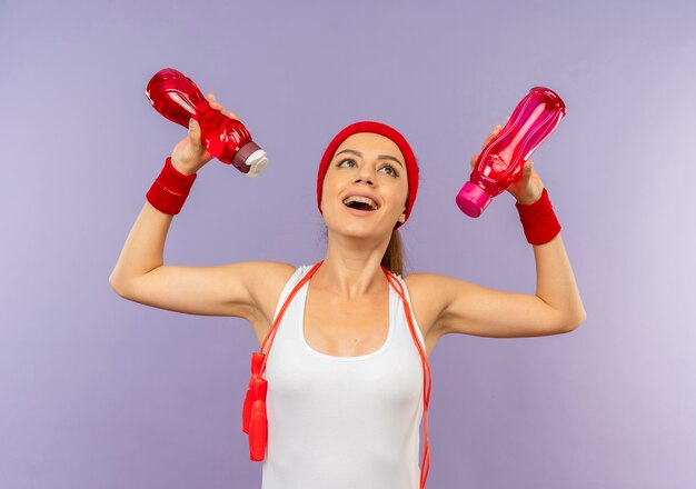 Молодая фитнес-женщина в спортивной одежде с повязкой на голову и скакалкой на шее держит бутылки с водой, глядя вверх счастливой и веселой, стоя над серой стеной
