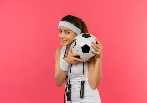 Young fitness woman in sportswear with headband and skipping rope around her neck holding soccer ball with smile on face standing over pink wall