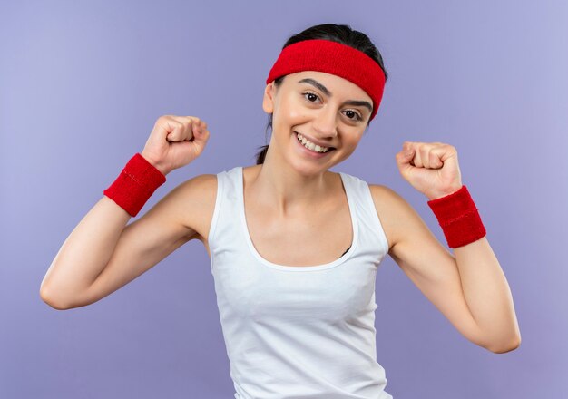 紫色の壁の上に幸せで前向きに立っている拳を元気に握り締めて笑っているヘッドバンドとスポーツウェアの若いフィットネス女性