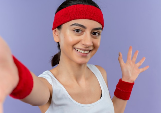 Молодая женщина фитнеса в спортивной одежде с повязкой на голову весело улыбается, делая приветственный жест руками, стоящими над фиолетовой стеной