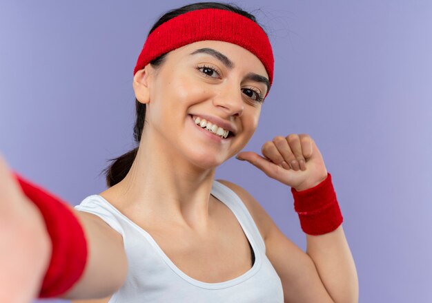 Молодая женщина фитнеса в спортивной одежде с повязкой на голову весело улыбается, делая приветственный жест руками, стоящими над фиолетовой стеной