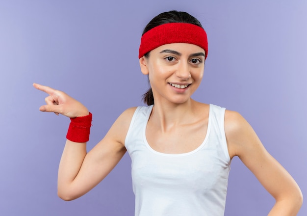 Young fitness woman in sportswear with headband smiling cheerfully pointing with index finger to the side standing over purple wall