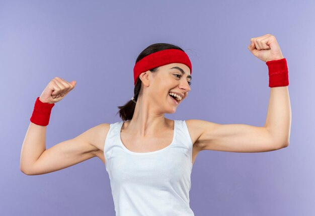 Young fitness woman in sportswear with headband smiling cheerfully raising fists happy and positive standing over purple wall