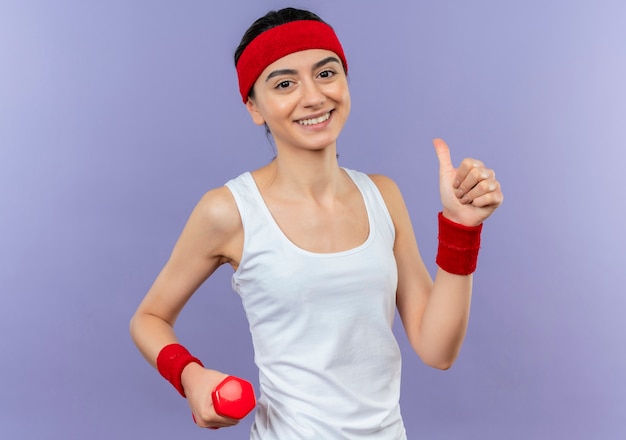 Молодая фитнес-женщина в спортивной одежде с повязкой на голову, весело улыбаясь, показывает палец вверх, стоя над фиолетовой стеной