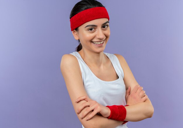 Young fitness woman in sportswear with headband smiling cheerfully with crossed arms on chest standing over purple wall