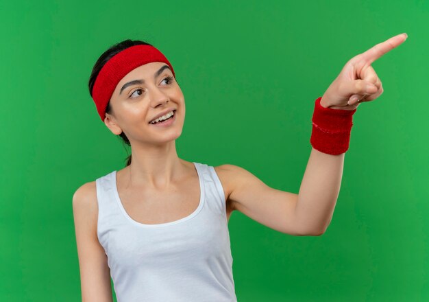 Young fitness woman in sportswear with headband smiling confident pointing with index finger to the side standing over green wall