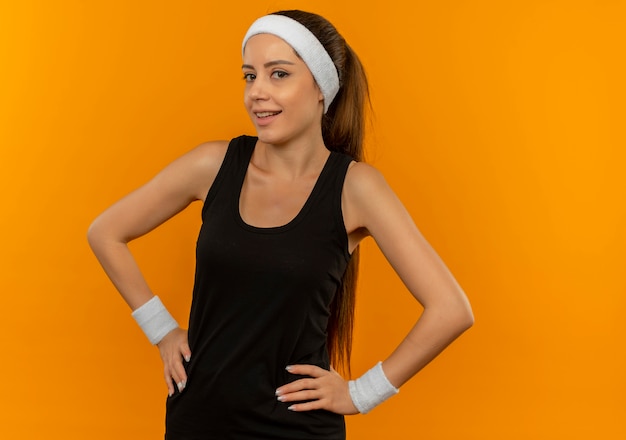 Young fitness woman in sportswear with headband smiling confident standing over orange wall