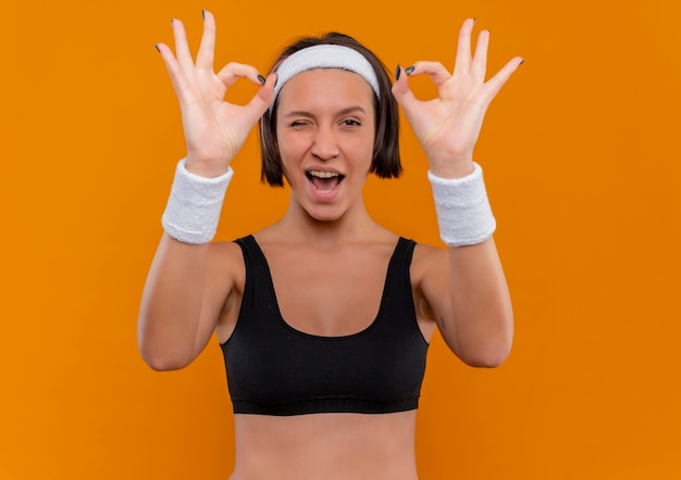 Young fitness woman in sportswear with headband smiling and winking making ok sign with both hands standing over orange wall