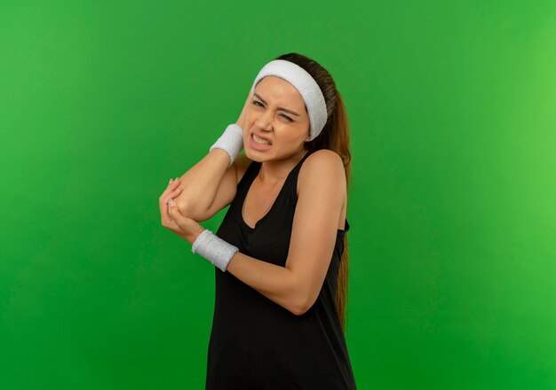 Young fitness woman in sportswear with headband touching elbow having pain standing over green wall