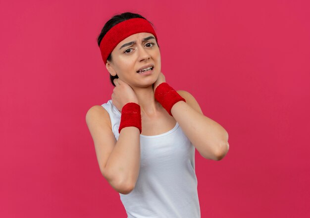 Young fitness woman in sportswear with headband touching her neck displeased feeling pain standing over pink wall