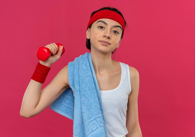 Молодая фитнес-женщина в спортивной одежде с повязкой на голову и полотенцем на плече, держа гантель в поднятой руке, выглядит уверенно, стоя над розовой стеной