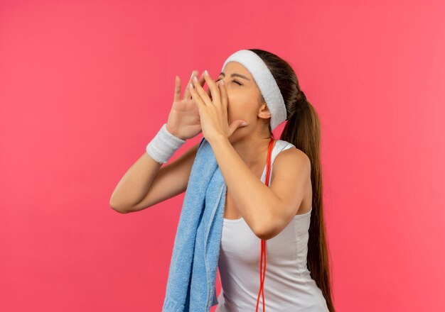 Young fitness woman in sportswear with headband and towel on her shoulder shouting with hands near mouth standing over pink wall