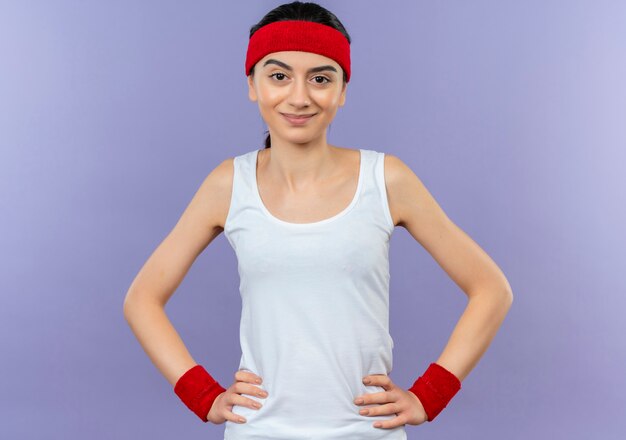 Young fitness woman in sportswear with headband with confident smile standing over purple wall