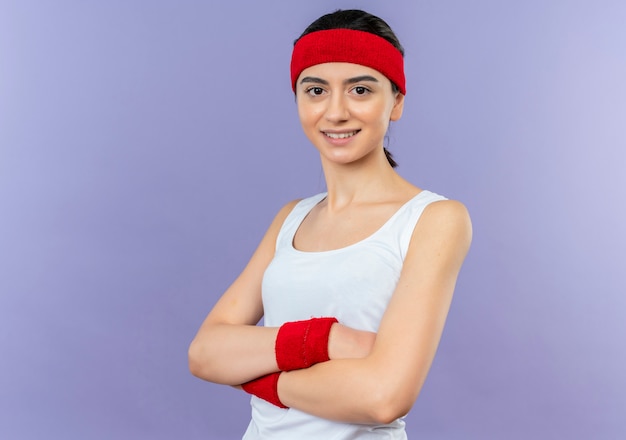 Giovane donna fitness in abiti sportivi con fascia con le braccia incrociate sul petto sorridente fiducioso in piedi sopra la parete viola