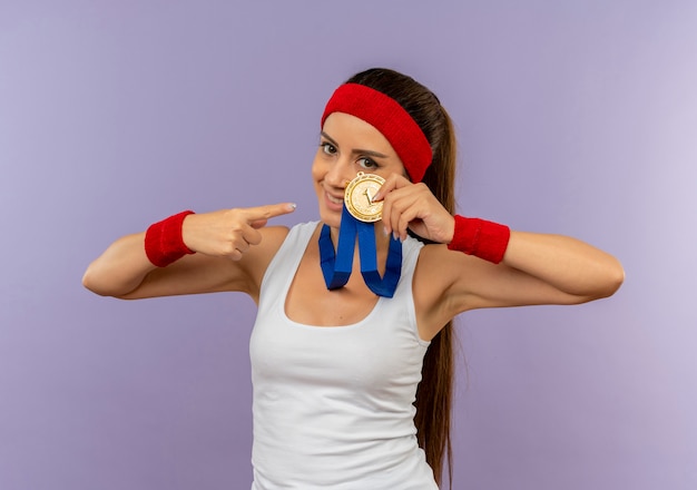 Free photo young fitness woman in sportswear with headband with gold medal around her neck showing and pointing with finger at it smiling standing over grey wall