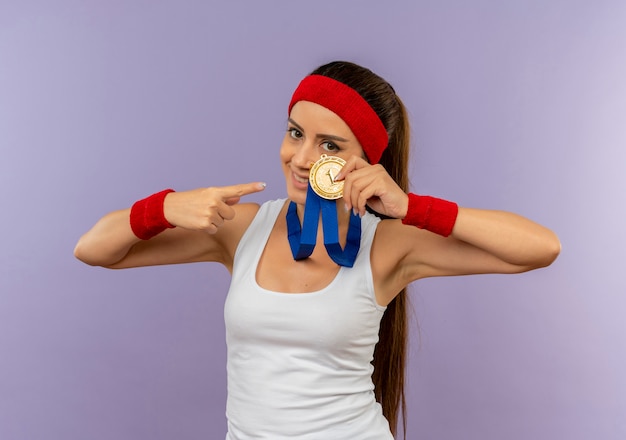 Молодая фитнес-женщина в спортивной одежде с повязкой на голову с золотой медалью на шее показывает и указывая пальцем на нее, улыбаясь, стоя над серой стеной