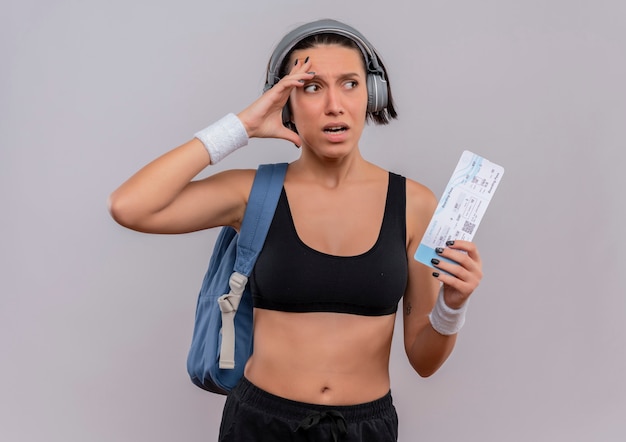 Young fitness woman in sportswear with headphones on head with backpack holding air ticket looking aside confused with fear expression standing over white wall