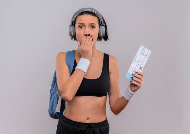 Young fitness woman in sportswear with headphones on head with backpack holding air ticket shocked covering mouth with hand standing over white wall