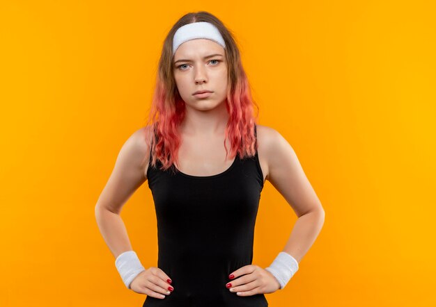 Молодая фитнес-женщина в спортивной одежде с серьезным уверенным выражением лица стоит над оранжевой стеной