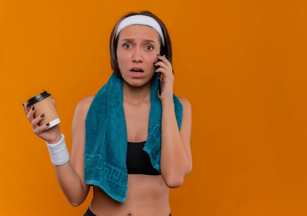 Young fitness woman in sportswear with towel on her neck holding coffee cup looking confused while talking on mobile phone standing over orange wall