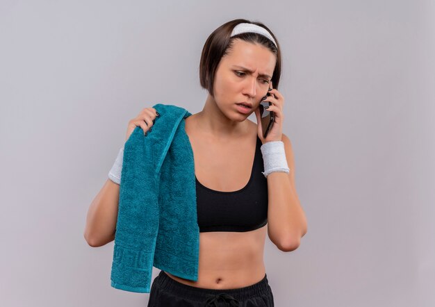 Young fitness woman in sportswear with towel on shoulder looking confused and anxious while talking on mobile phone standing over white wall