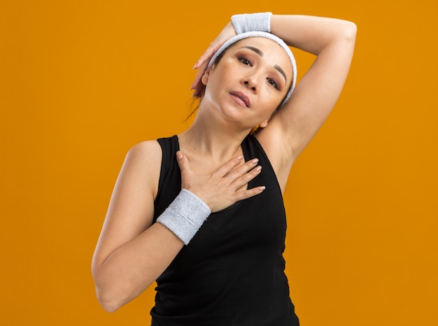 Бесплатное фото Молодая фитнес-женщина с повязкой на голову и повязками чувствует дискомфорт при прикосновении к ее голове и груди, стоя над оранжевой стеной