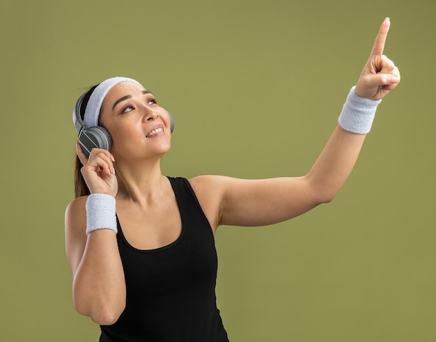 Бесплатное фото Молодая фитнес-женщина с повязкой на голову и наушниками, наслаждаясь своей любимой музыкой, указывая указательным пальцем вверх, улыбаясь, стоя над зеленой стеной