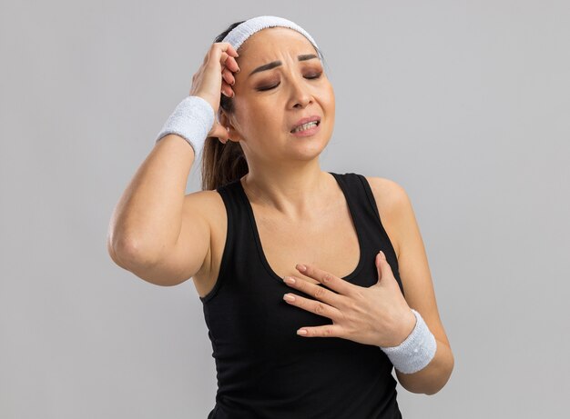 Young fitness woman with headband and armbands feeling unwell touching her head standing over white wall