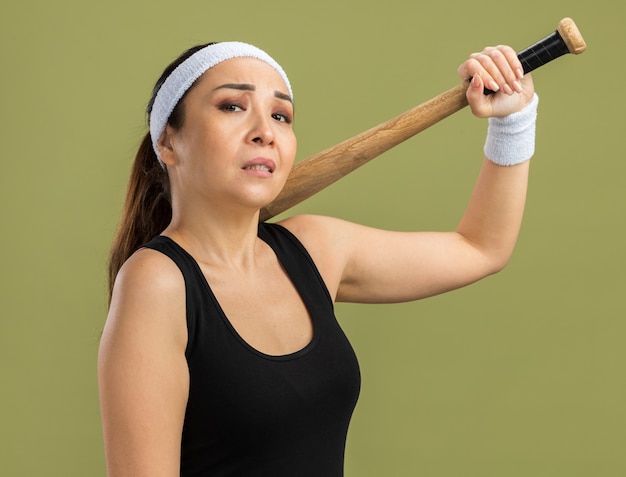 Free photo young fitness woman with headband and armbands holding baseball bat   with confident expression