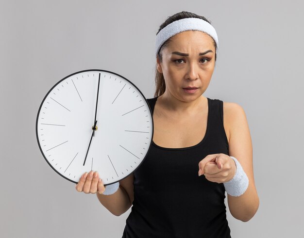 Young fitness woman with headband and armbands holding wall clock pointing with index finger  being angry standing over white wall