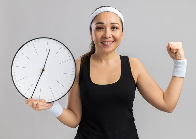 Молодая фитнес-женщина с повязкой на голову и нарукавными повязками, держащая настенные часы с улыбкой на лице, сжимая кулак, стоя над белой стеной