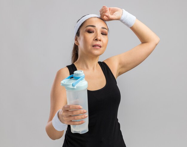 Young fitness woman with headband and armbands holding water bottle looking tired standing over white wall