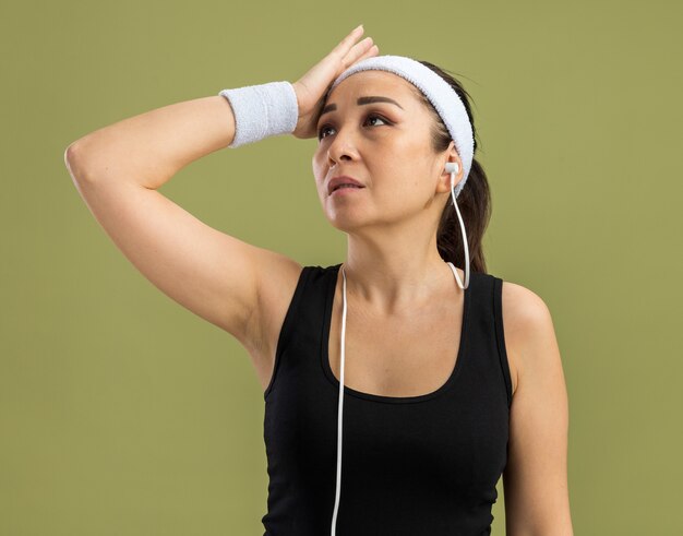 Молодая фитнес-женщина с повязкой на голову и нарукавными повязками смотрит в сторону, озадаченная, положив руку на голову, стоя над зеленой стеной