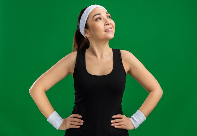 Young fitness woman with headband and armbands looking aside smiling confident with arms at hip  