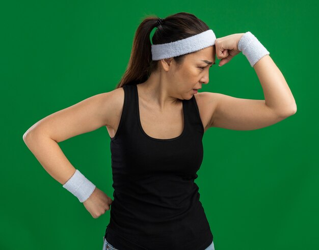 Free photo young fitness woman with headband and armbands looking aside with pensive expression thinking standing over green wall