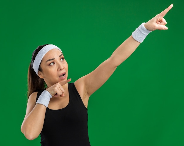 Free photo young fitness woman with headband and armbands looking aside with serious face pointing with index fingers up standing over green wall