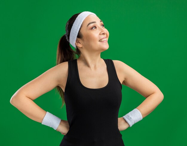 Young fitness woman with headband and armbands looking aside with smile on face with arms at hip  