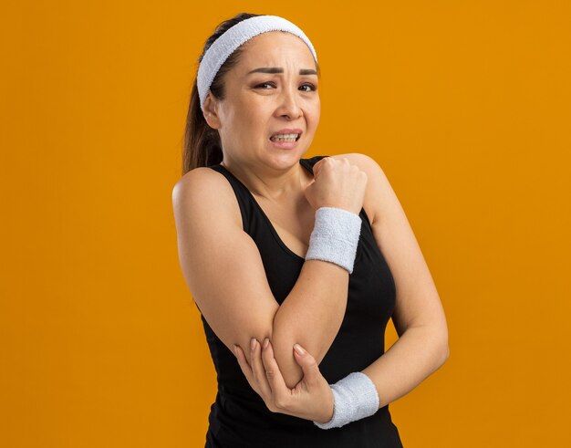 Young fitness woman with headband and armbands looking unwell touching her elbow feeling pain  