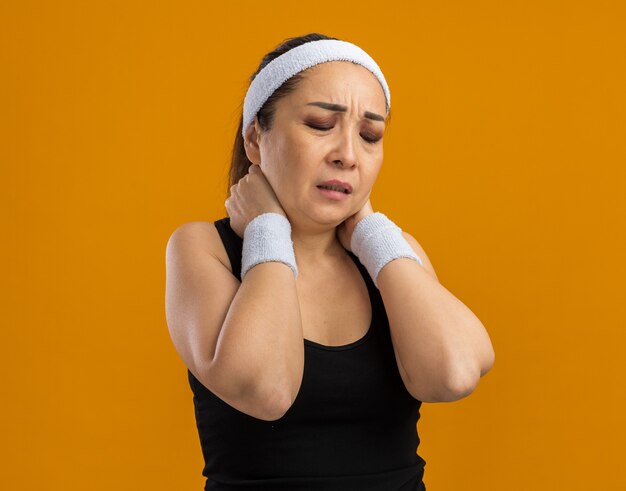 Молодая фитнес-женщина с повязкой на голову и нарукавными повязками выглядит нездоровой, касаясь ее шеи, чувствуя боль, стоя над оранжевой стеной