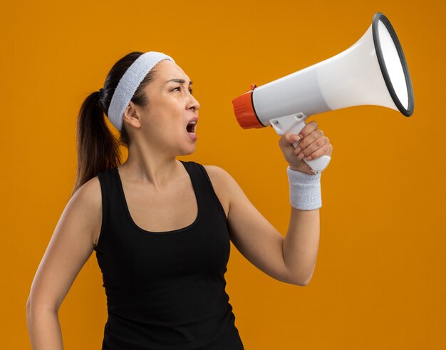 Молодая фитнес-женщина с повязкой на голову и нарукавными повязками кричит в мегафон с агрессивным выражением лица