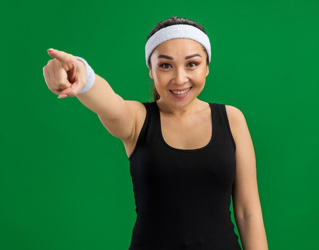 Молодая фитнес-женщина с повязкой на голову и нарукавными повязками, уверенно улыбаясь, указывая указательным пальцем, стоя над зеленой стеной