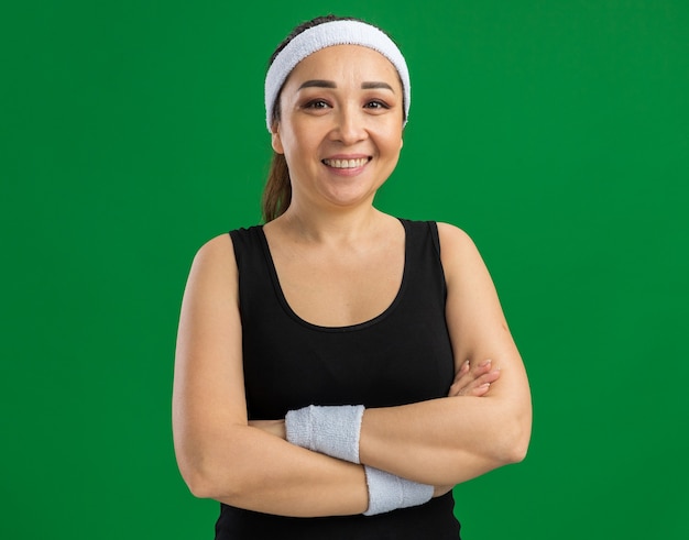 Giovane donna fitness con fascia e bracciali sorridente sicura di sé con le braccia incrociate in piedi sul muro verde green