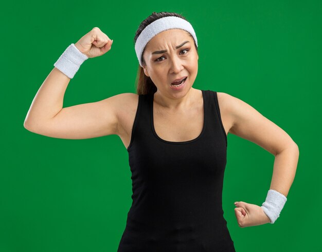 Молодая фитнес-женщина с повязкой на голову и нарукавными повязками напряженно и уверенно поднимает кулаки, показывая силу и мощь, стоя над зеленой стеной