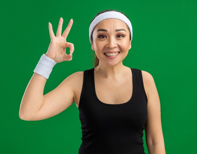 Молодая фитнес-женщина с повязкой на голову и нарукавными повязками с улыбкой на лице делает знак ОК