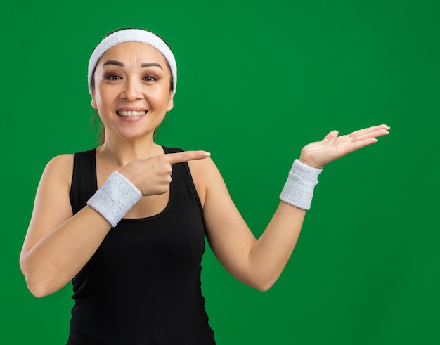 Young fitness woman with headband and armbands   with smile on face presenting with arm of hand copy sapce pointing with index finger to the side  